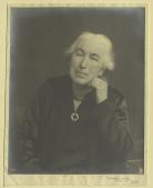 Photograph of Clemence Housman in old age, with white hair, a black dress and brooch, and a dark dress. Her chin rests on her left hand and she is looking directly at the camera. 