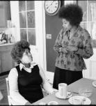 Toni Morrison and Alice Walker are seen in a kitchen. Toni is sitting to the left looking up at Alice, standing to the right.