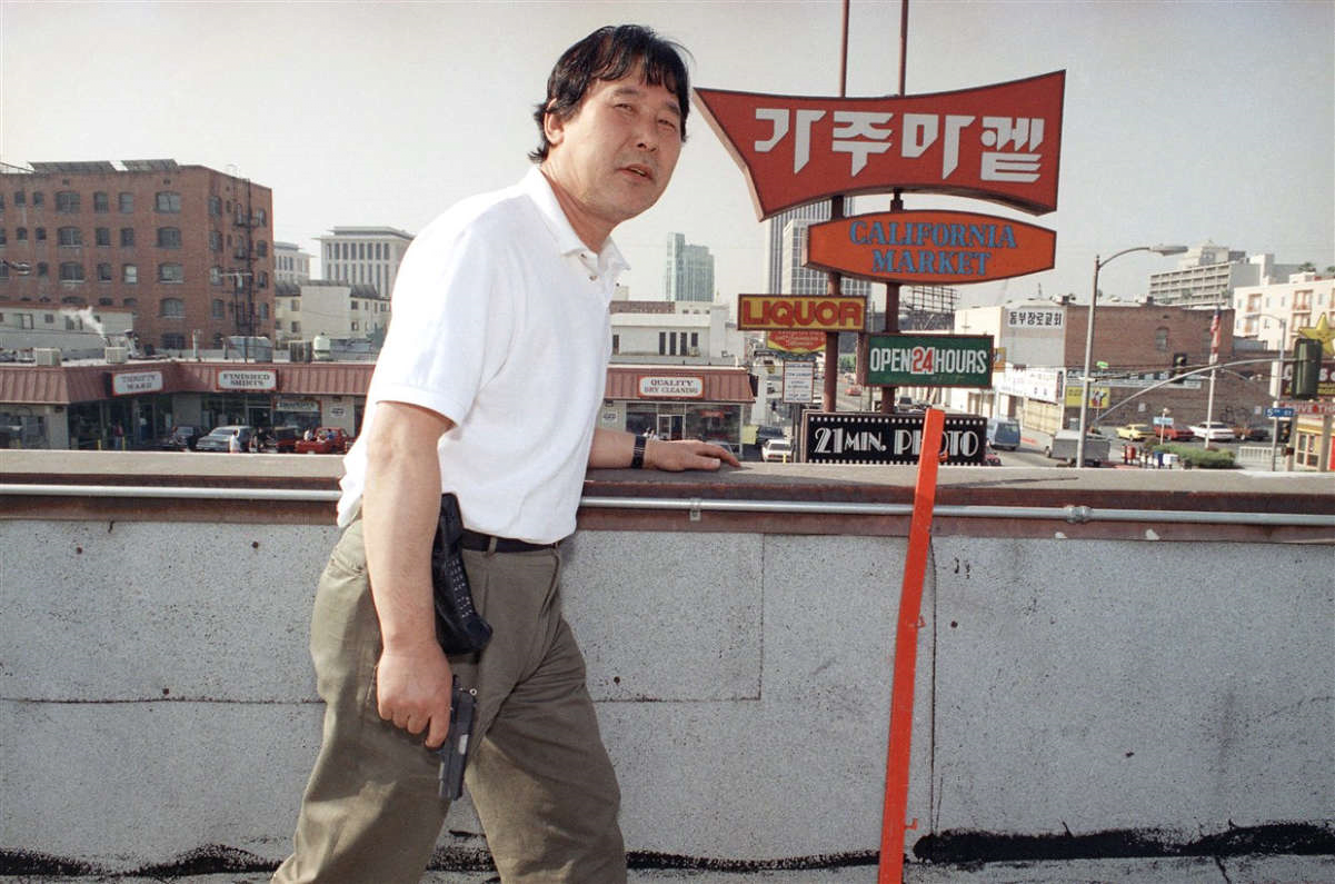 This image depicts a Korean man defending his store from a rooftop in Los Angeles. Furthermore, he is armed with a handgun (Lah).