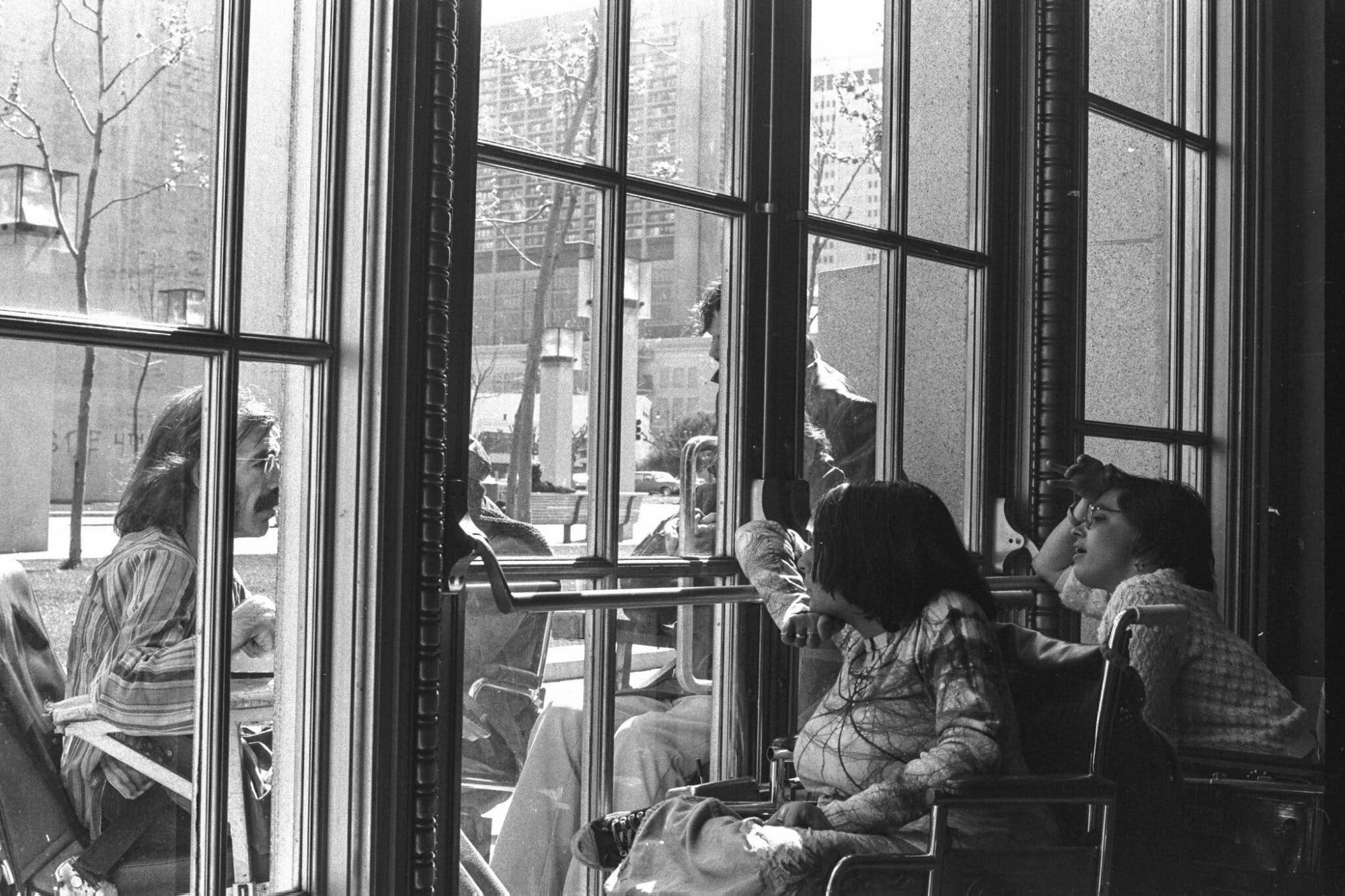 People in wheelchairs inside a building looking out of a glass door, where a man with a wheelchair looks in.