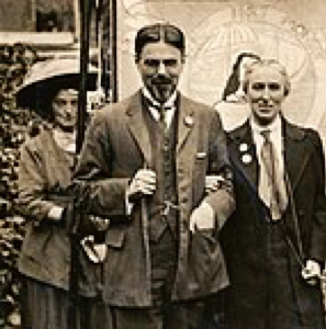 Photograph, printed, paper, monochrome, Laurence and Clemence Housman wearing suffrage badges and standing in front of a suffrage banner. LSE LIbrary. Wikimedia Commons